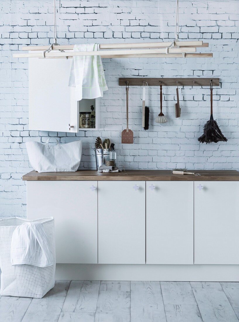 White base units, washing-up brushes hung from hooks and clothes airer suspended from ceiling in utility room with brick-patterned wallpaper