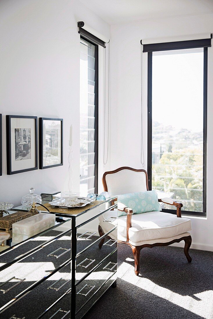 Antique armchair in corner between black-framed, tall narrow windows next to mirrored chests of drawers