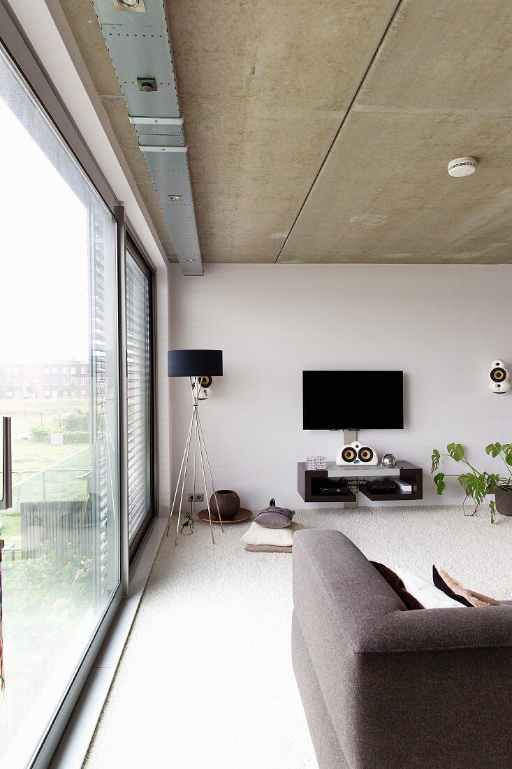 Zeitgenössisches Wohnzimmer mit Fensterfront und Sichtbetondecke, im Hintergrund Stehleuchte mit schwarzem Schirm neben Flachbildschirm an Wand