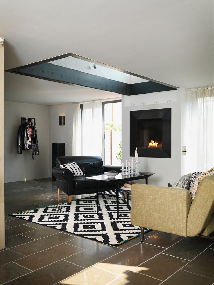 Black leather armchair and sofa around coffee table on black and white patterned rug in living room with skylight