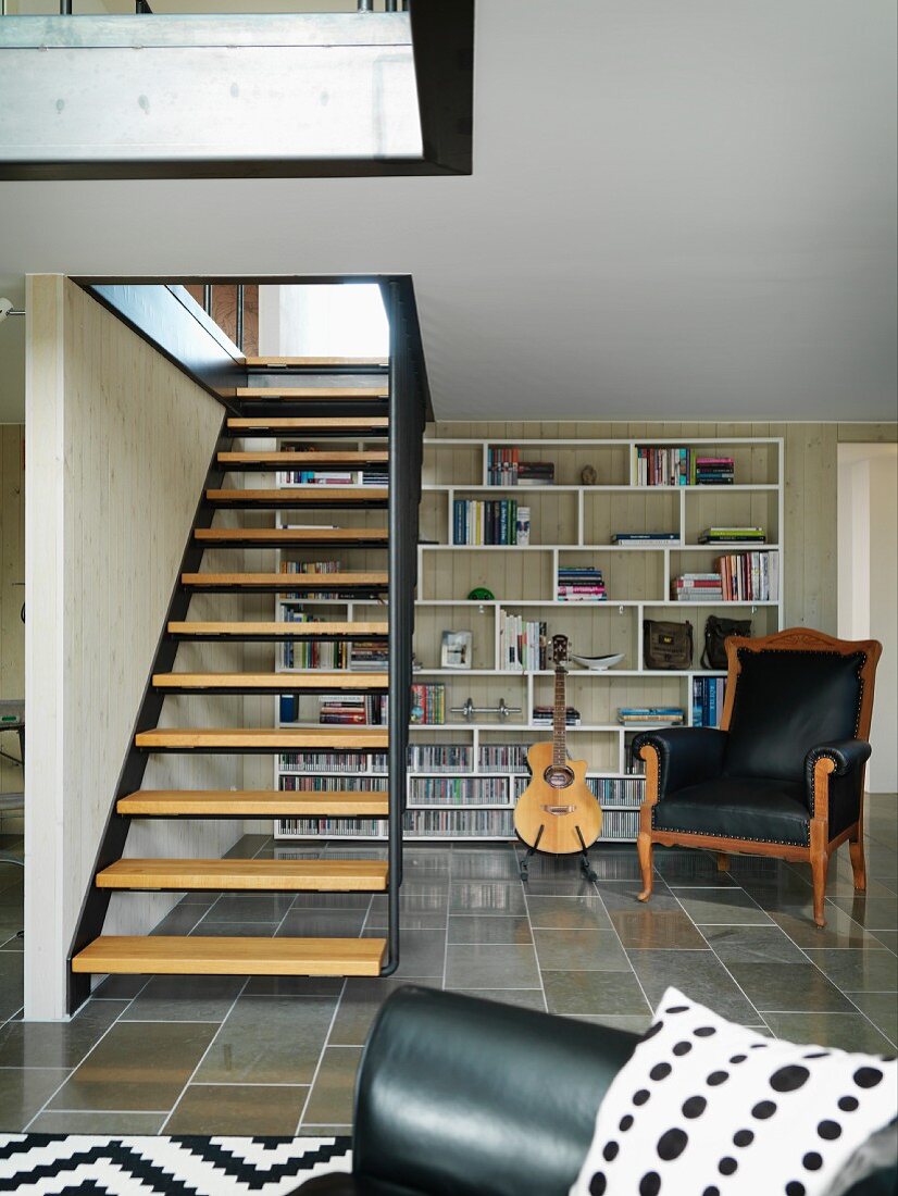 Metal staircase with wooden treads in living room with black leather armchair in front of modern shelving in background