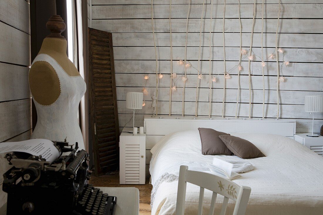 Simple bed with wooden headboard made of branches decorated with fairy lights, vintage typewriter and tailors' dummy in white, wood-clad bedroom