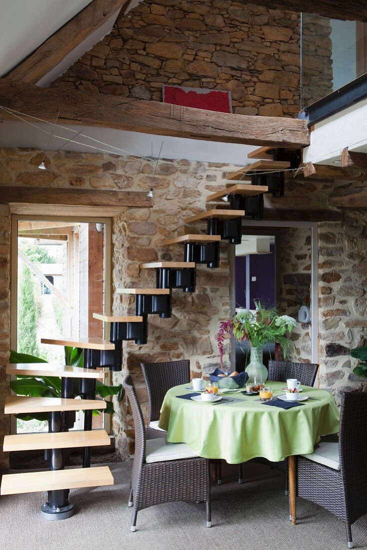 Table with green tablecloth and wicker chairs at foot of window staircase in interior with stone walls