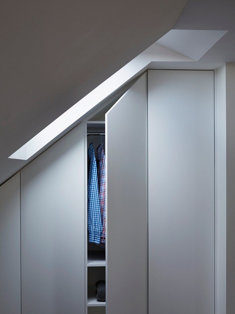 Wardrobe without handles and with one half-open door fitted below sloping ceiling; light falling through skylight