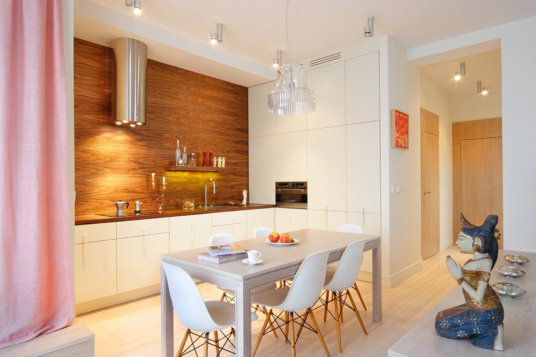 Dining table and Eames Plastic Chairs in front of white and wooden fitted kitchen with stainless steel extractor hood; ethnic figurine in foreground