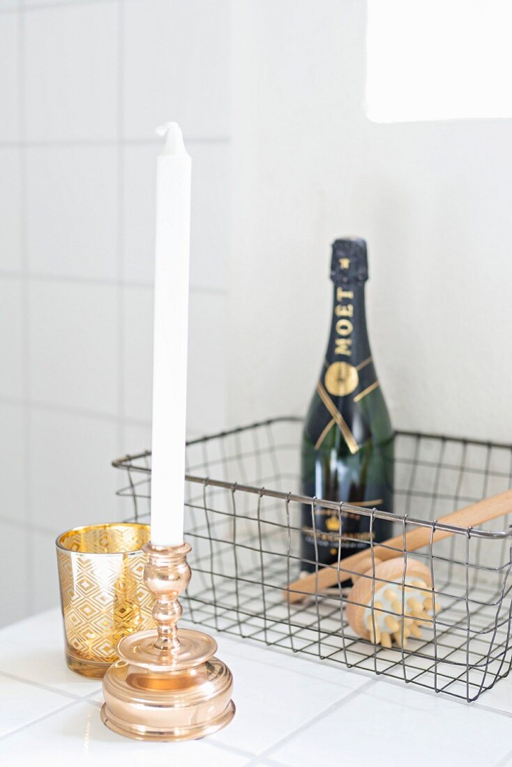 Goldfarbener Kerzenhalter mit weisser Kerze und Windlicht vor Maschendrahtkorb mit Champagnerflasche