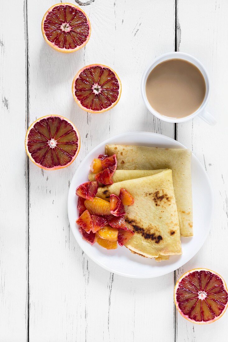 Crepes with blood oranges on a plate with a cup of coffee and halved blood oranges next to it