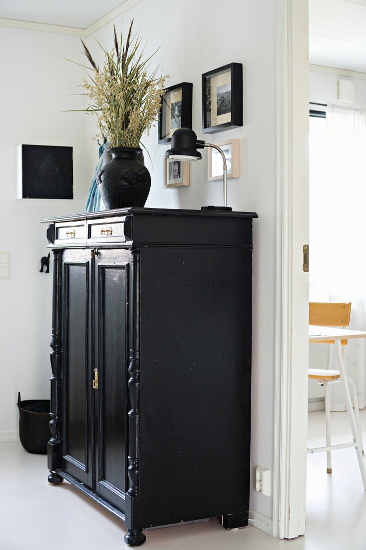 Vase of dried flowers and table lamp on top of black-painted cupboard with carved door