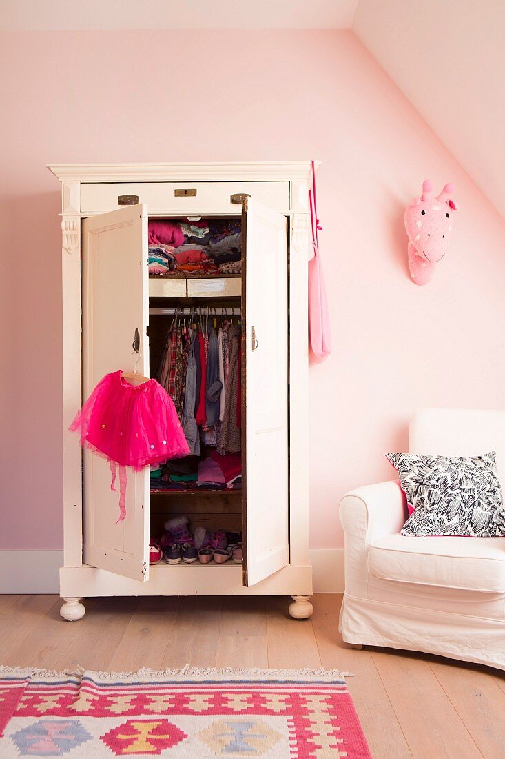 Kleiderschrank mit offenen Türen und Blick auf Kinderkleidung, seitlich weisser Polstersessel vor rosa getönter Wand