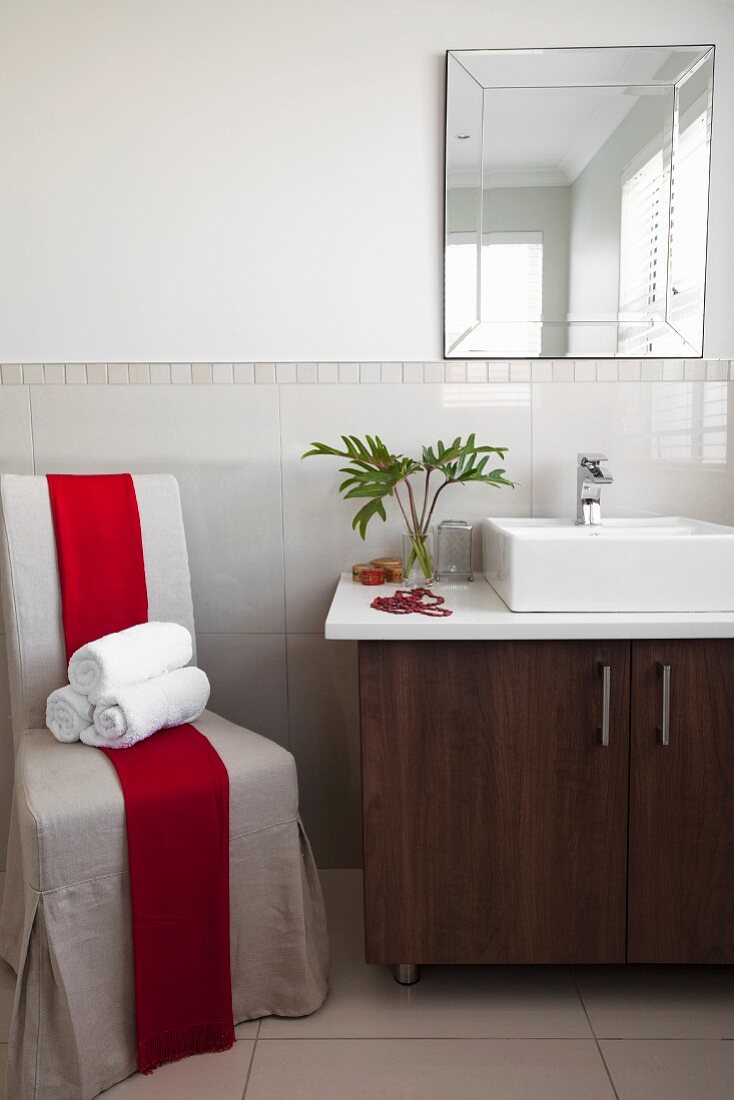 Rolled towels on loose-covered chair with red sash next to washstand with base cabinets and countertop basin