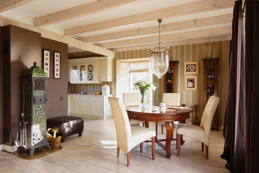 Essplatz mit Stilmöbeln unter Hängeleuchte mit grossem Glasschirm an Holzbalkendecke, in elegantem, offenem Wohnraum, im Hintergrund weiße Landhausküche