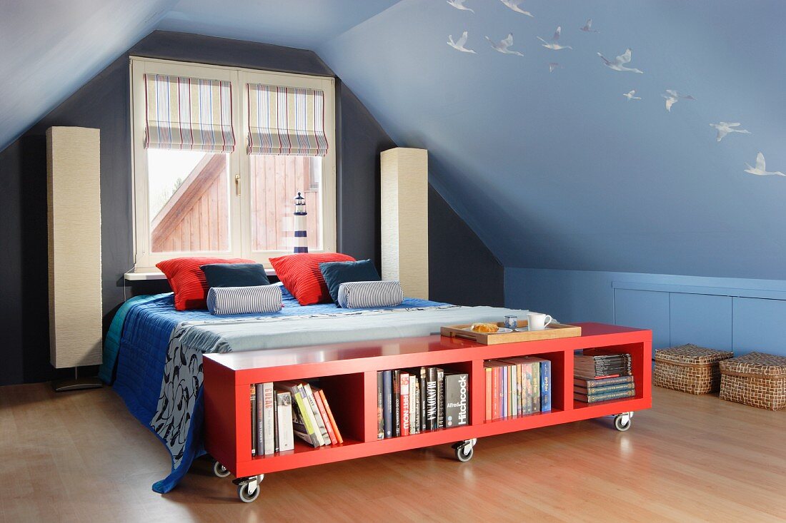 Low, red sideboard on castors at foot of bed and standard lamps against gable end wall in attic room painted mid blue