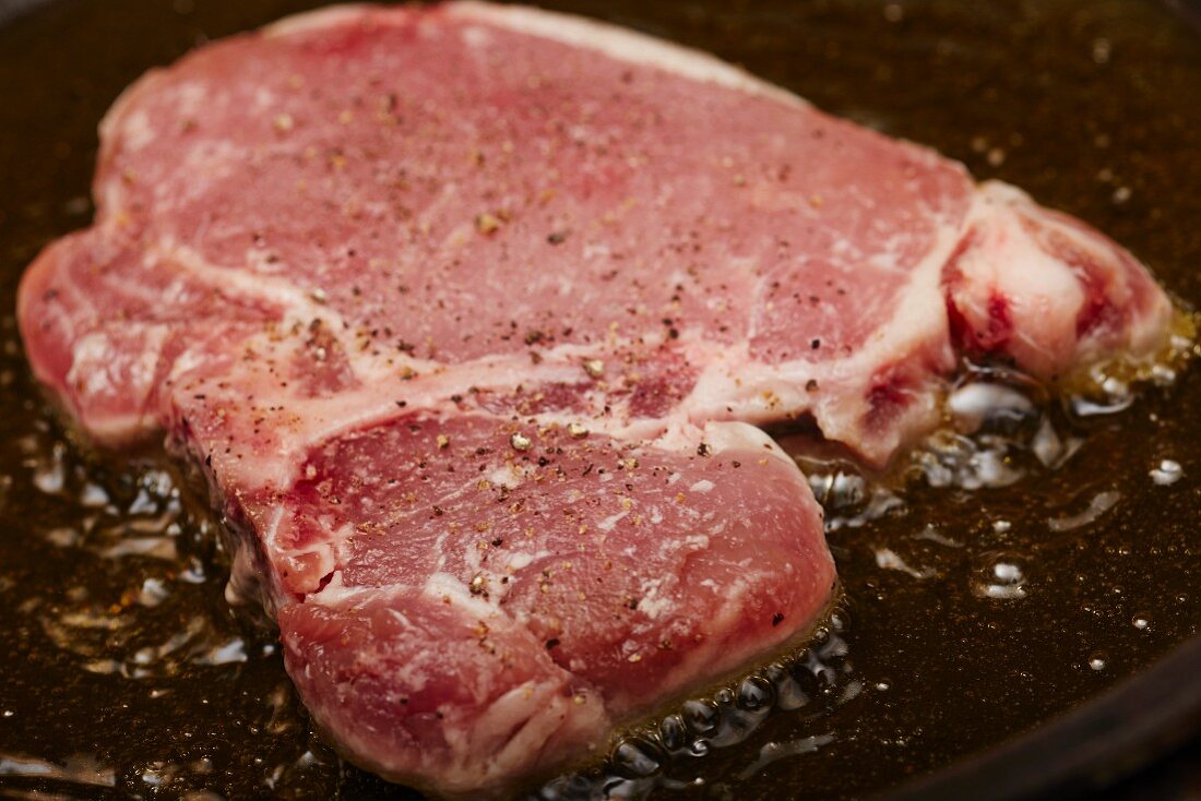 A pork chop seasoned with salt and pepper being fried in olive oil