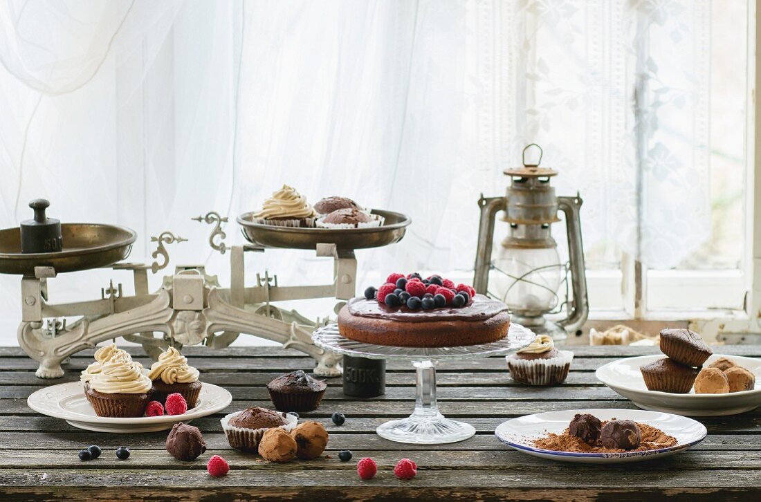 Cupcakes, Pralinen und Beerenkuchen mit Vintage-Waage auf altem Holztisch vor dem Fenster