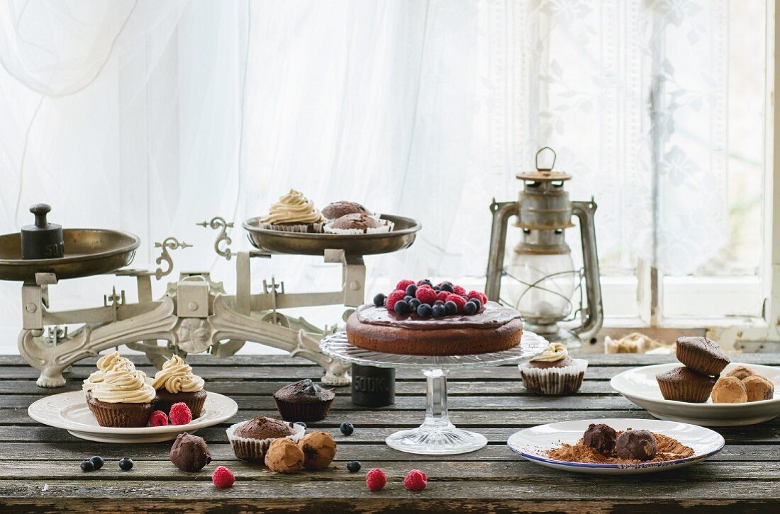 Cupcakes, pralines and berry cake with vintage scales on an old wooden table in front of a window