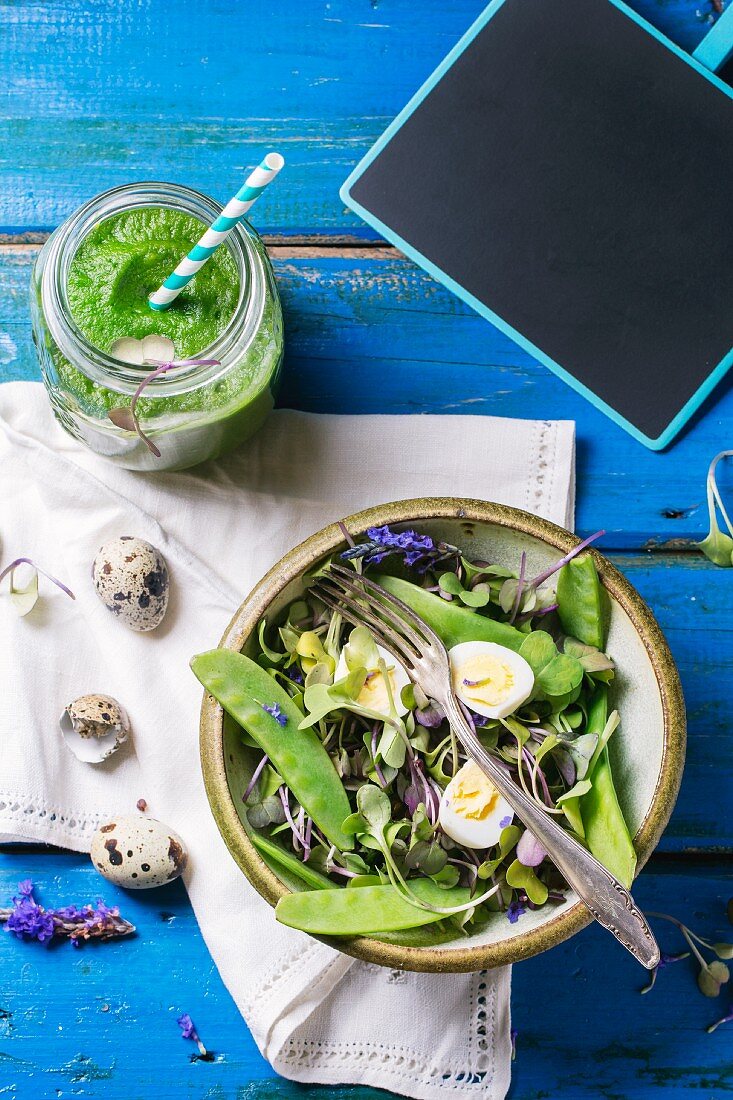 A mixed leaf salad with young peas and quail's eggs served with a herb smoothie on a blue wooden table
