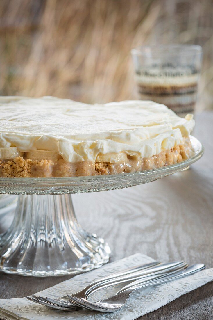 Tarte mit Bananen, Karamell und Mascarpone auf Kuchenständer, im Hintergrund ein Glas Latte Macchiato