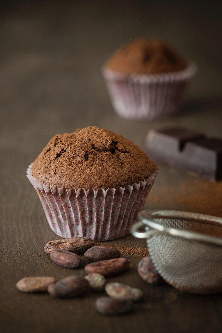 Schokoladenmuffins mit Kakaopulver und Kakaobohnen auf Tischplatte mit Sieb und Kuvertüre