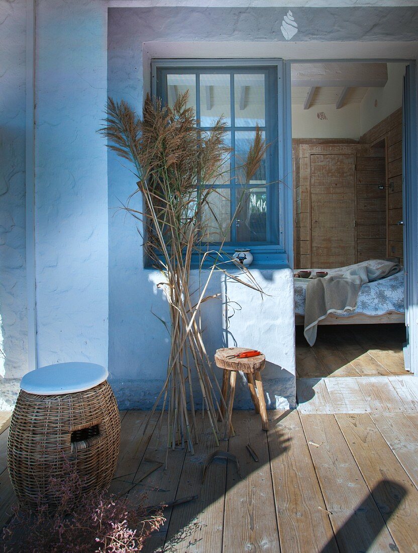 Wicker stool with seat cushions and bunch of pampas grass on wooden terrace with view into bedroom