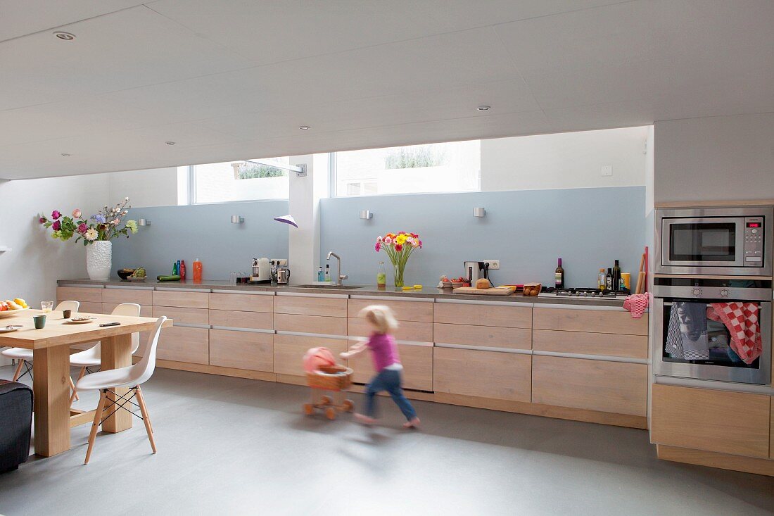 Long kitchen counter with pale wooden doors and pale blue splashback in kitchen-dining room; little girl playing