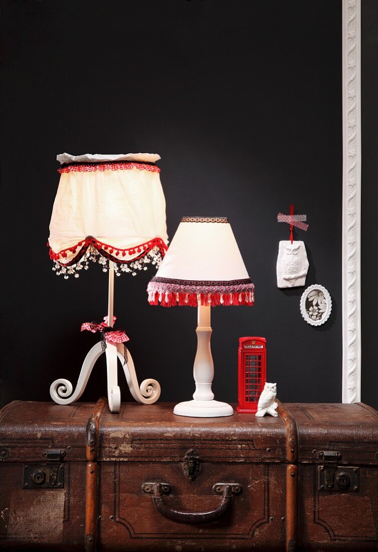 Lampshades decorated with pompoms, fringes and lace trim on country-house-style bases on top of antique steamer trunk
