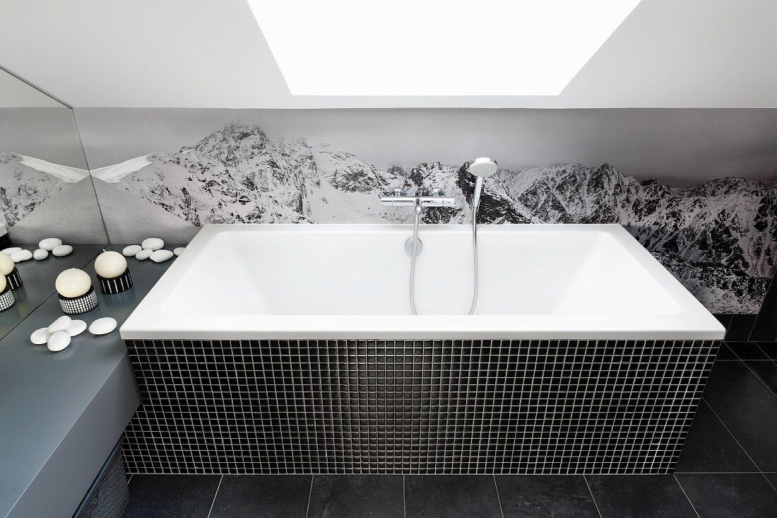 Bathtub with black mosaic-tiled surround against photo mural of mountains below skylight