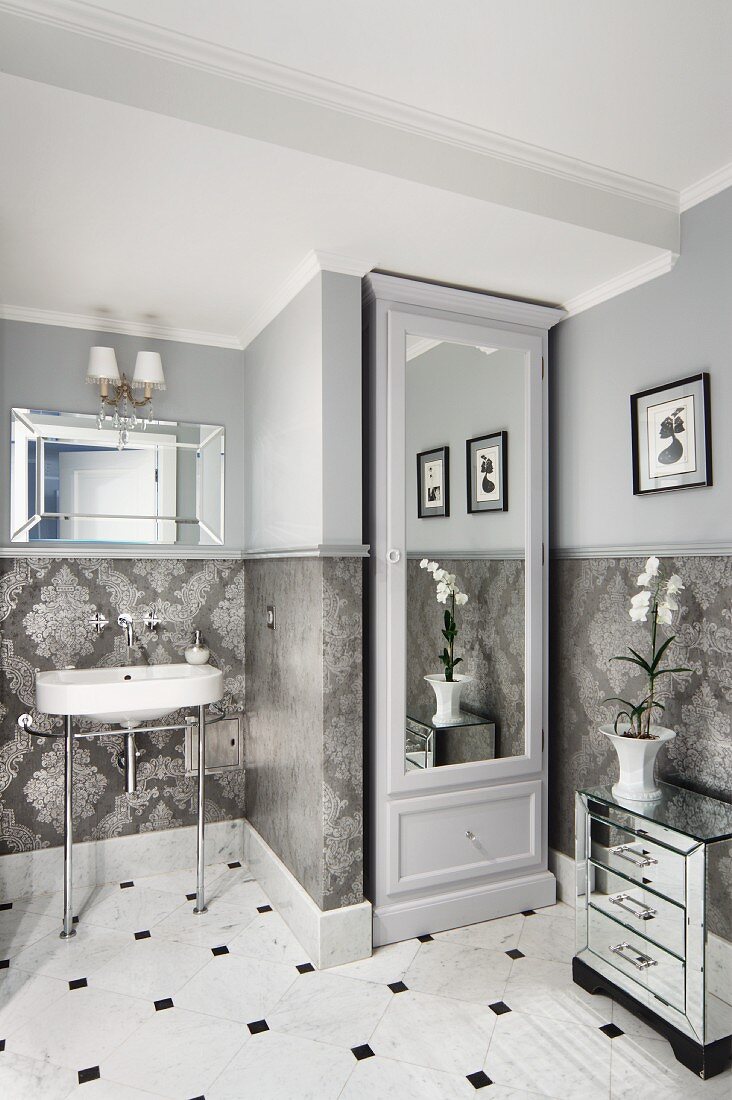 Mirrored cupboard in niche next to retro washstand with metal frame in traditional bathroom with walls tiled below dado rail and painted pale grey above