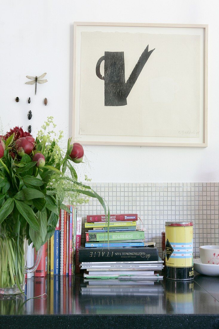 Stacked books on worksurface against tiled splashback below minimalist artwork on wall