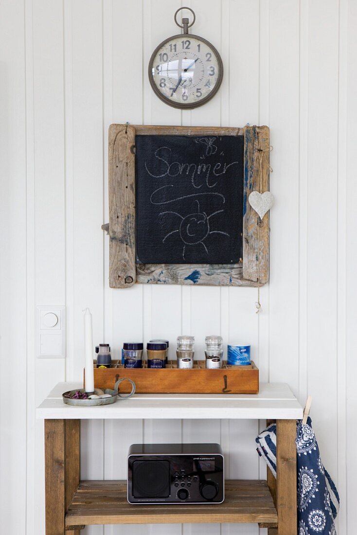 Box of spice jars on wooden shelving unit with white top below blackboard and station clock on white wooden wall