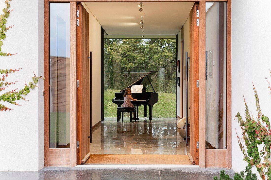 View into foyer with girl playing grand piano and glass wall with view of garden
