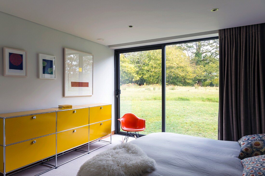 Bedroom with yellow, steel-tube sideboard next to orange, classic shell chair next to glass wall with view of landscaped garden