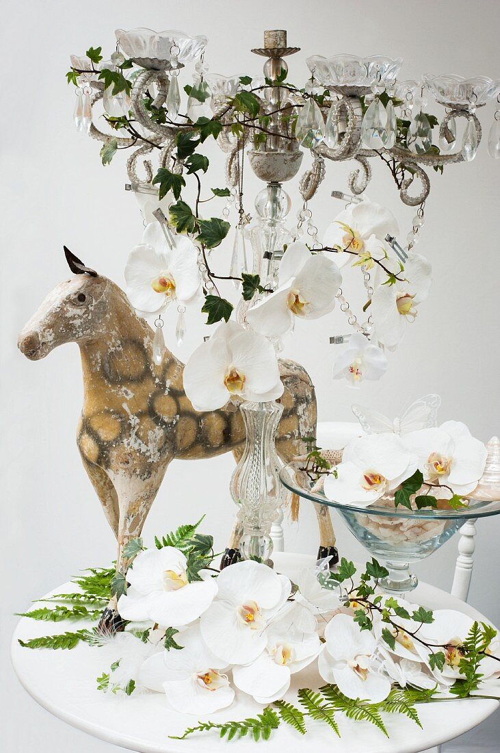 weiße Orchideenblüten in Schale und auf Kerzenständer mit Glasschmuck, dahinter antikes Miniatur Pferd auf weißem Tisch