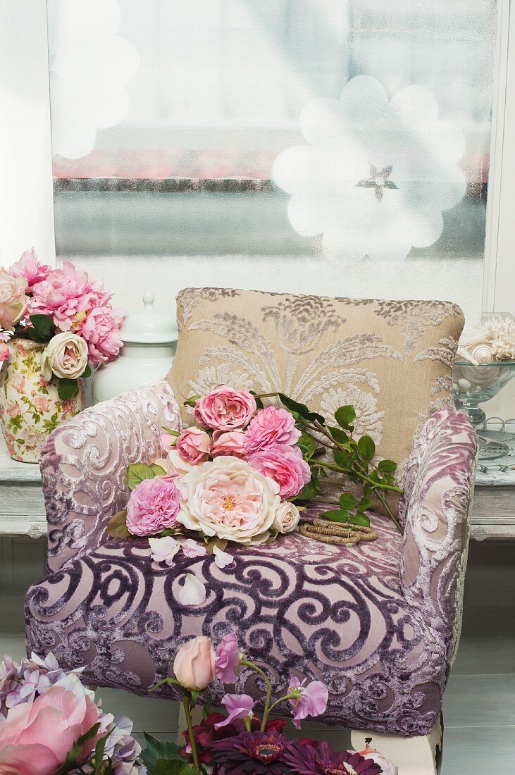 Peonies on armchair with a burnt out pattern cover against a wall hung with a picture