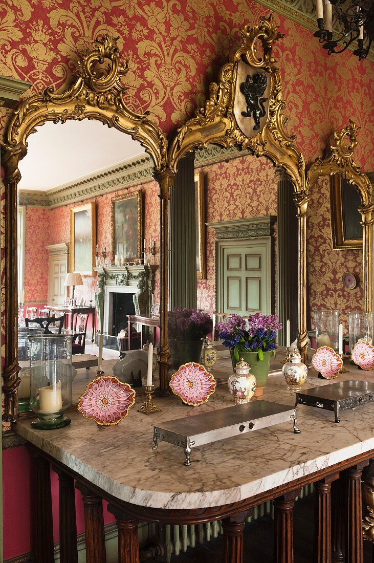 Gilt-framed triple mirror on sideboard with marble top in dining room with red and gold patterned wallpaper