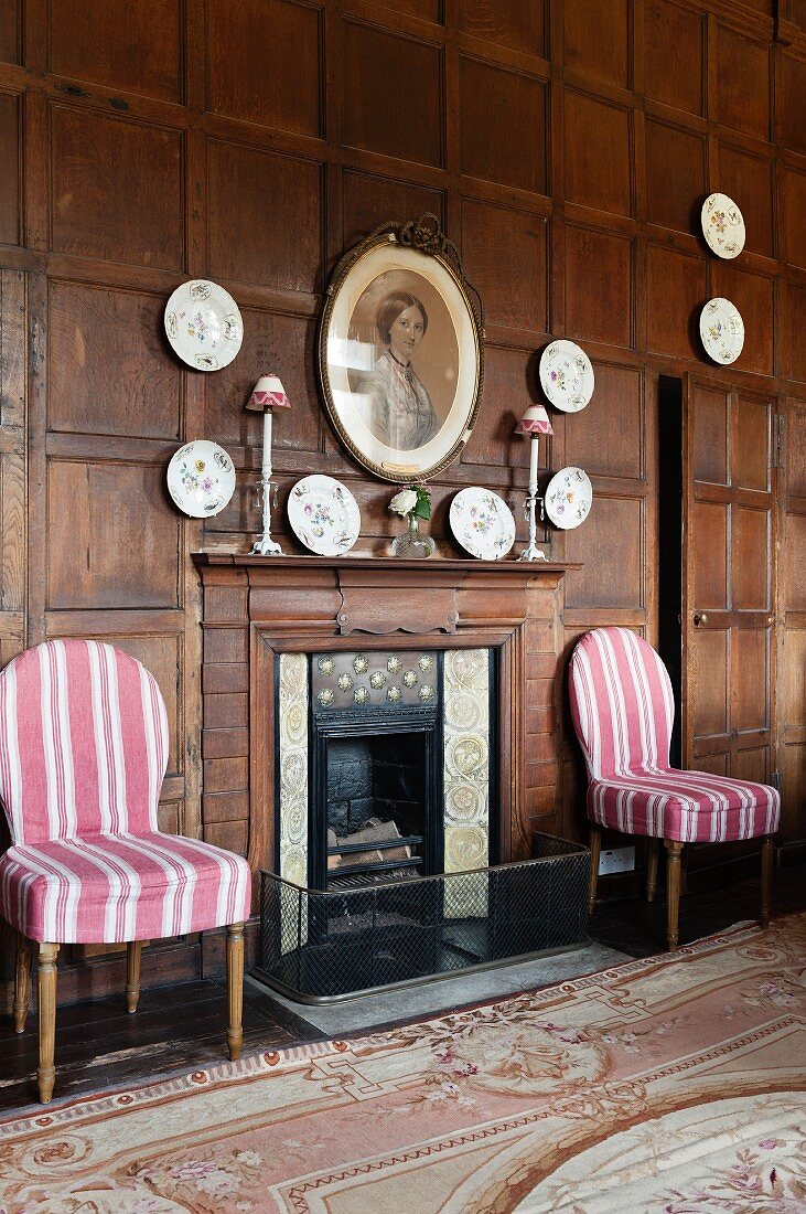 Open fireplace flanked by white and pink striped chairs, decorative china wall plates on wood-panelled wall and hand-made Aubusson-style rug