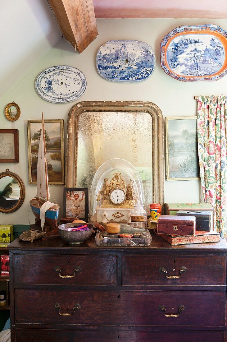 Table clock and vintage mirror on top of wooden chest of drawers below decorative plates on wall