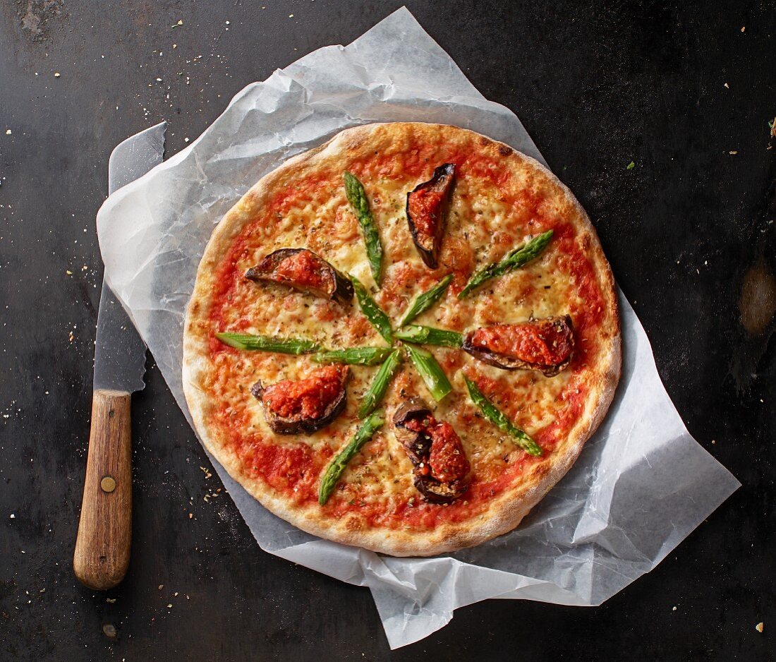 Pizza Margherita with asparagus and aubergines on a piece of paper with a knife