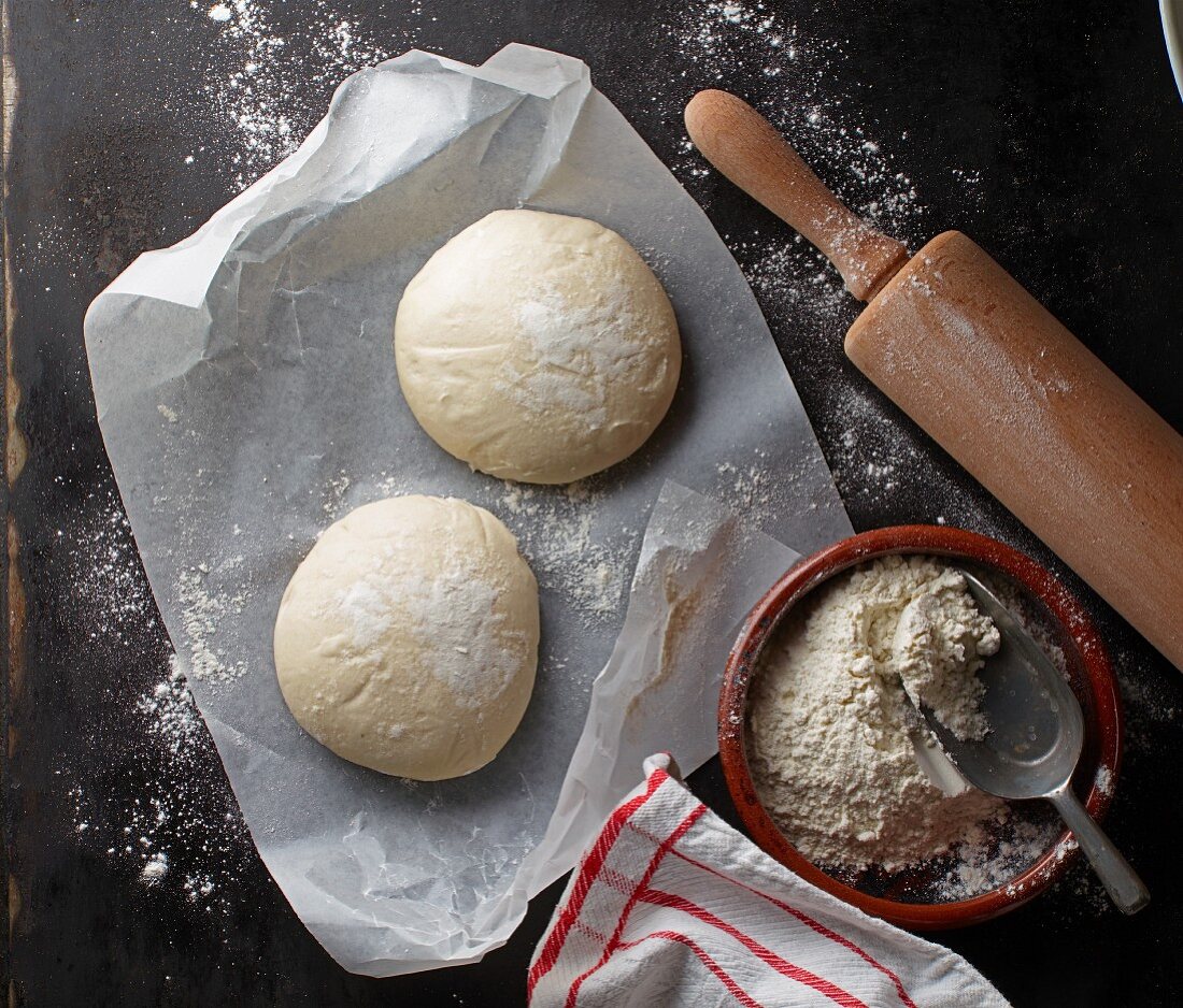 Two balls of pizza dough on a piece of paper with flour and a rolling pin