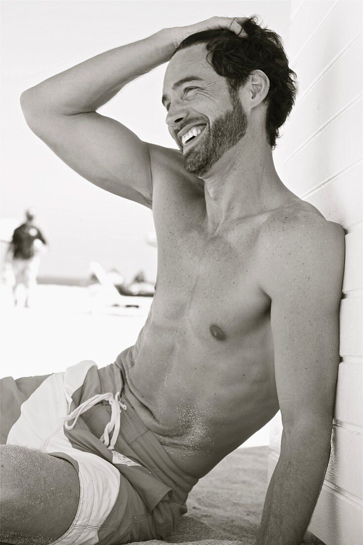 A laughing young man sitting on a beach wearing swimming shorts (black-and-white shot)