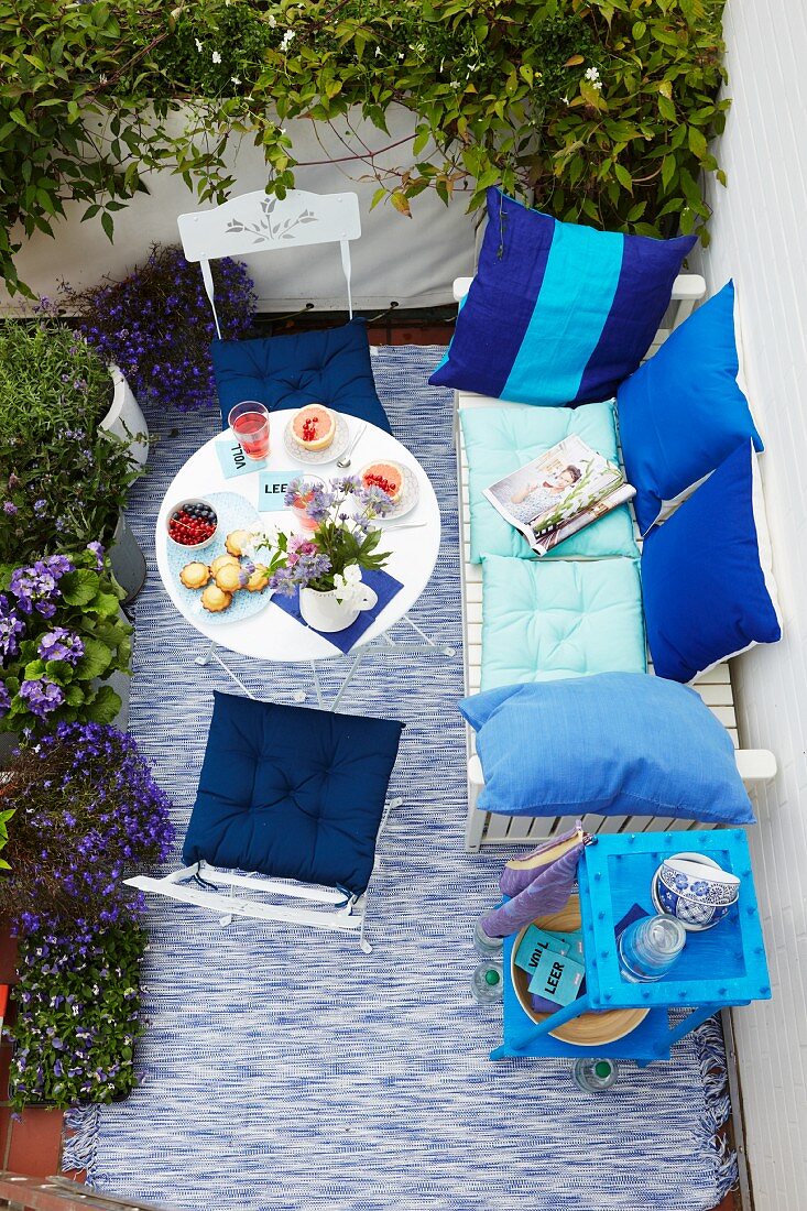 A summery balcony seen from above with a laid table and aqua coloured cushions on white, outdoor furniture