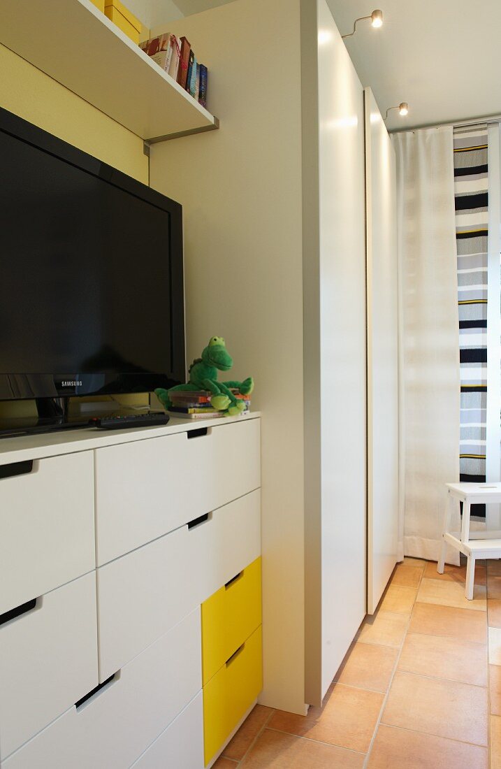 Weisses Schlafraum-Interieur mit gelben Akzenten; Fernseher auf Kommode neben dem Kleiderschrank