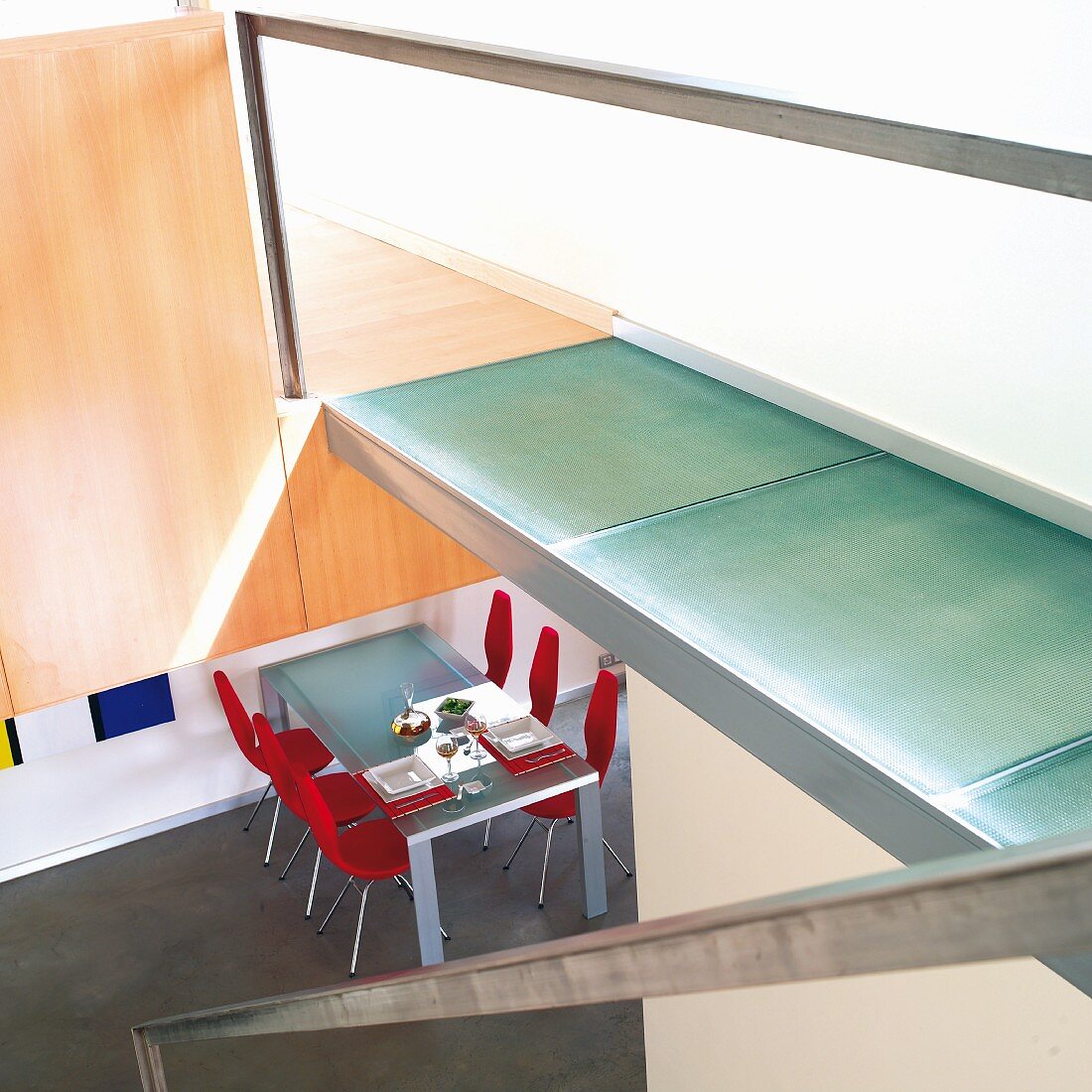 Staircase with walkway landing made from glass panels, view into open-plan dining area with table and red chairs