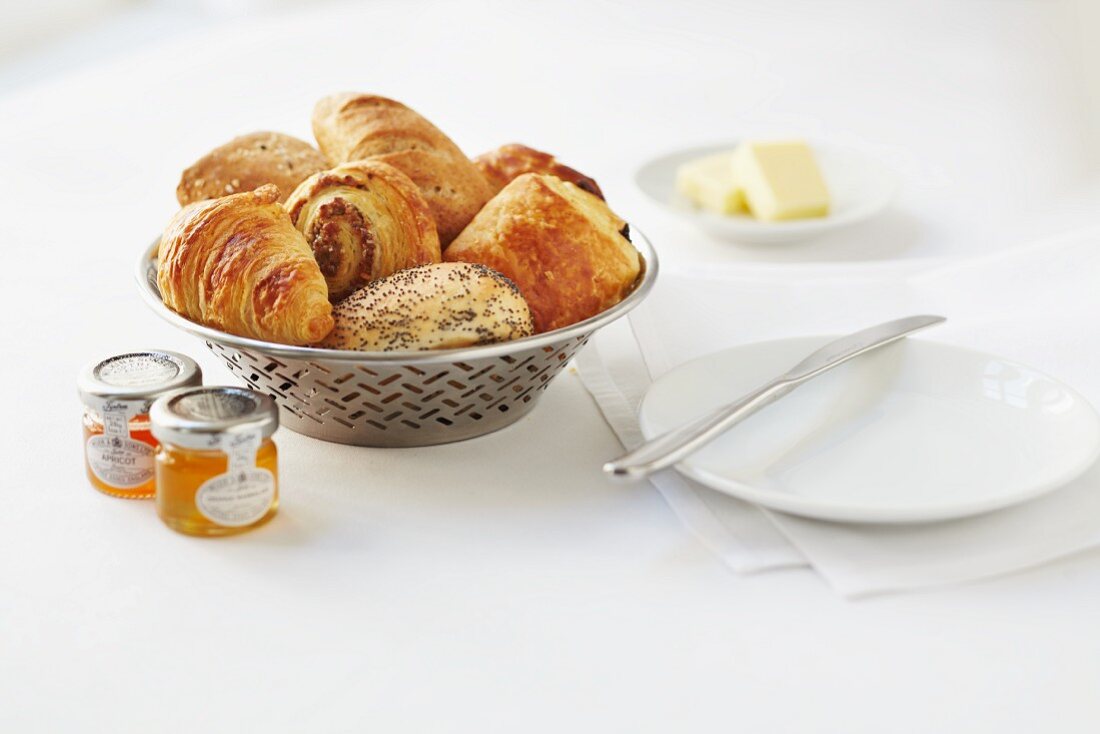 Frühstücksbrötchen, Croissants, Plundergebäck und Mohnbrötchen im Brotkorb, Marmeladengläser und Butter