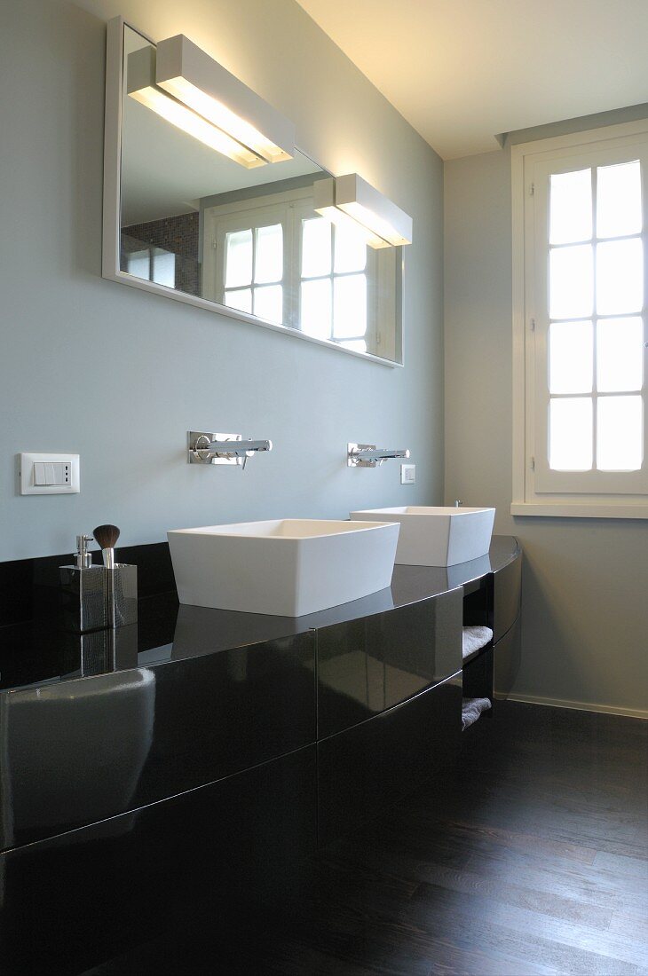 Schwarzer, gebogener Waschtisch mit weissen Aufbauschüsseln und Wandarmatur, darüber Spiegel mit Beleuchtung an grau getönter Wand