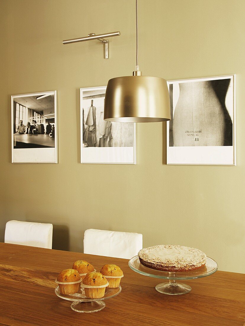 Muffins und Kuchen auf Esstisch; Pendelleuchte mit Metallschirm und künstlerische Schwarzweissfotos an der Wand