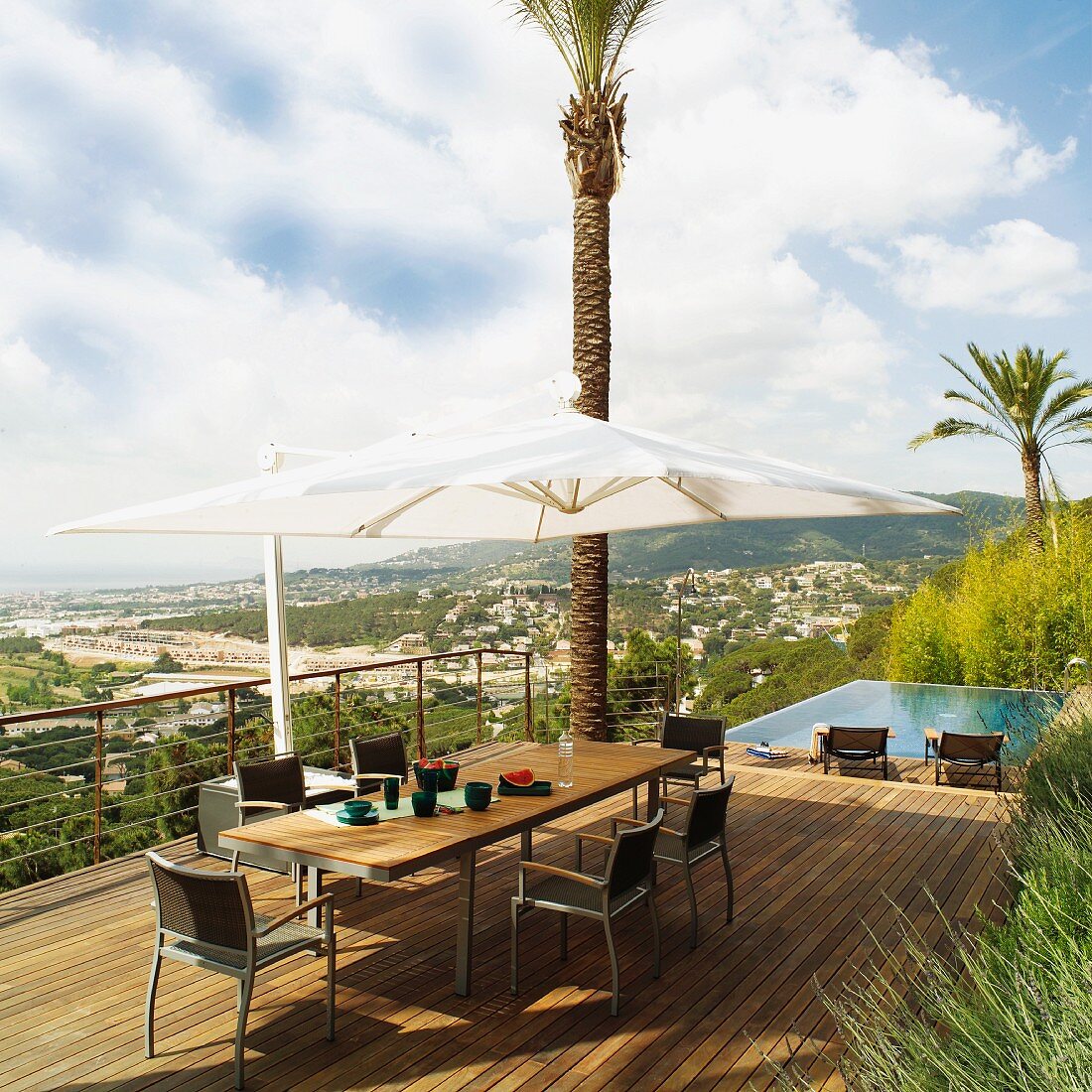 Dining set and parasol on wooden deck with view of town on hillside; infinity pool in background