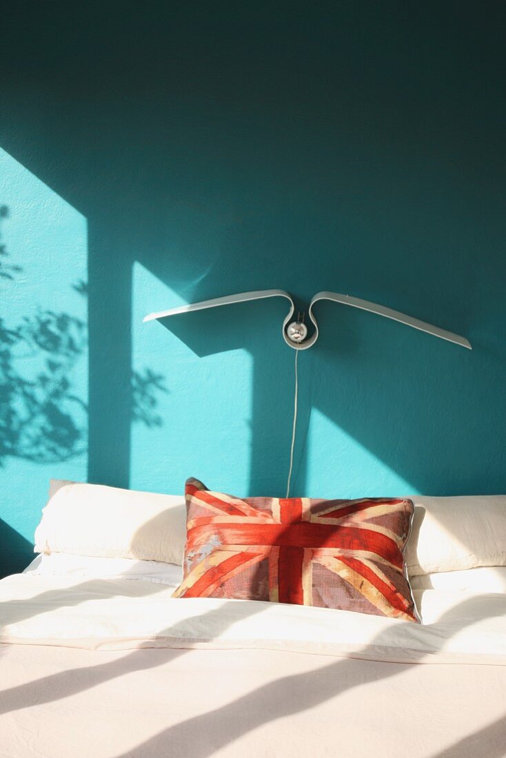 Union Jack Kissen auf Doppelbett vor türkis getönter Wand, darüber eine stilisierte Möwensilhouette als Wandleuchte