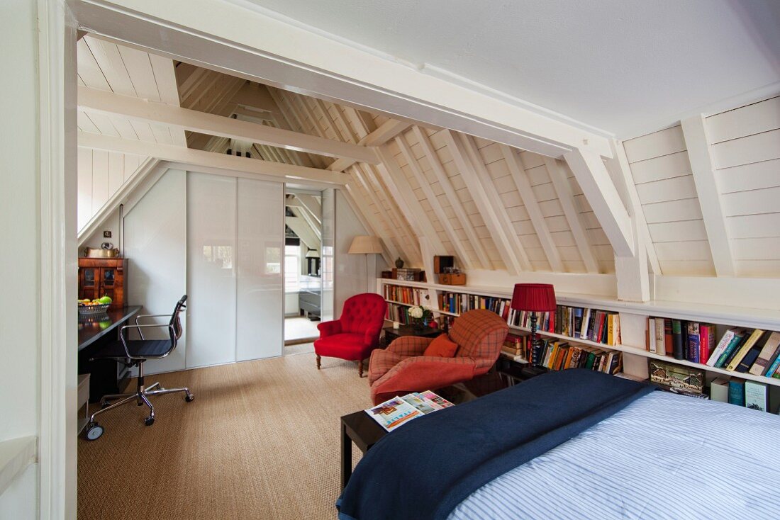 Multifunctional attic room with home office, seating area, bookcases and bed under exposed, white-painted wooden roof structure