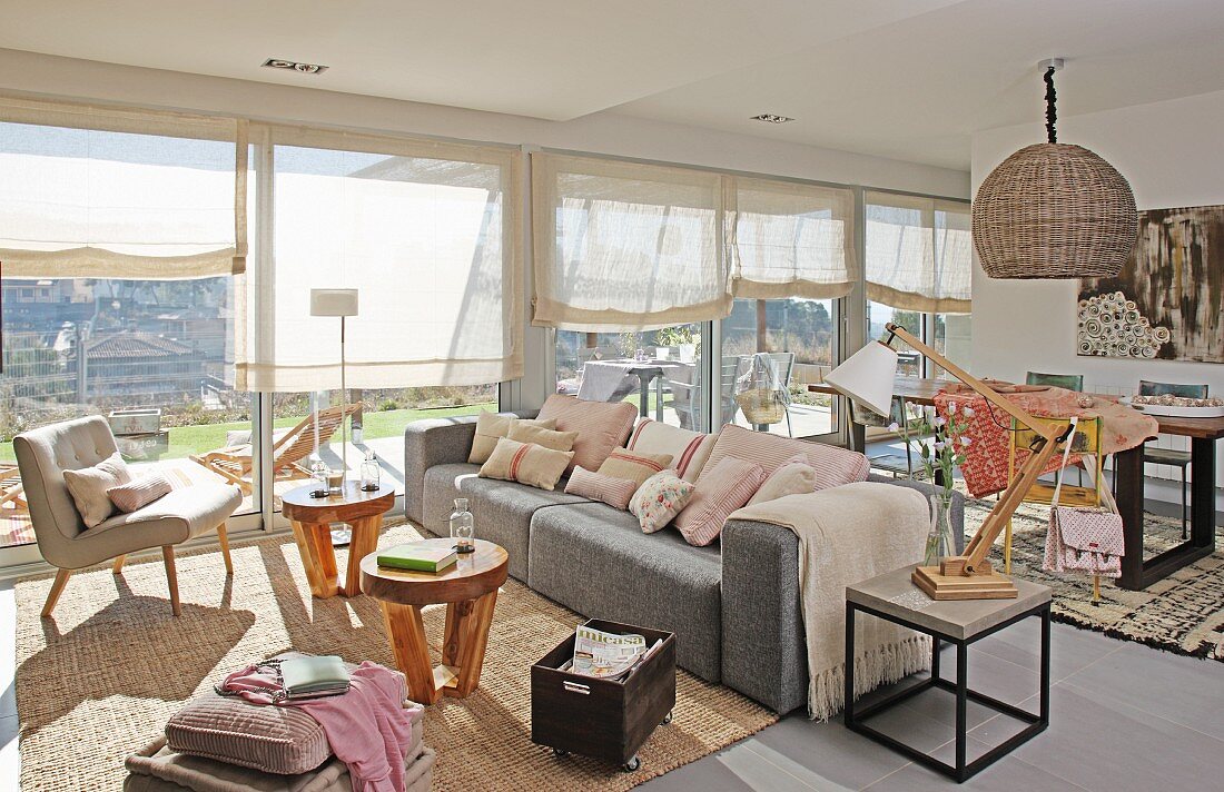 Sonnendurchfluteter Wohnraum, im Loungebereich Polstersessel und hellgraues Sofa vor kleinen Holz Beistelltischen, im Hintergrund Essplatz