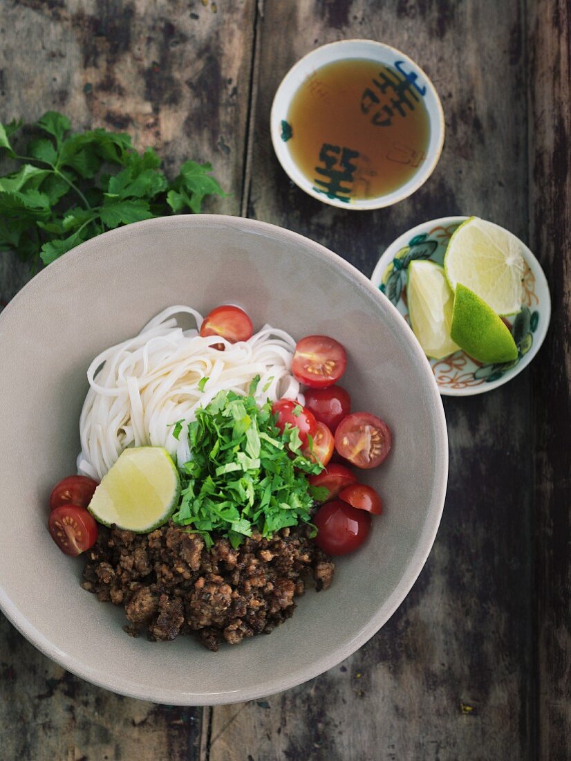 Rice noodles with fried minced meat, tomatoes and coriander in a porcelain bowl on a wooden table